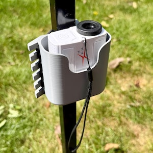 May include: A light gray plastic holder mounted to a black vertical post, containing a white laser rangefinder with a black eyepiece and a red star-shaped logo. The rangefinder features a black wrist strap hanging down from the side of the device.