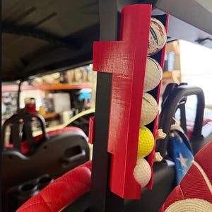 May include: A red golf ball holder attached to a black support post, holding four golf balls. The golf balls are white, yellow, and patterned. The holder is mounted on a golf cart, with a red seat visible in the foreground.