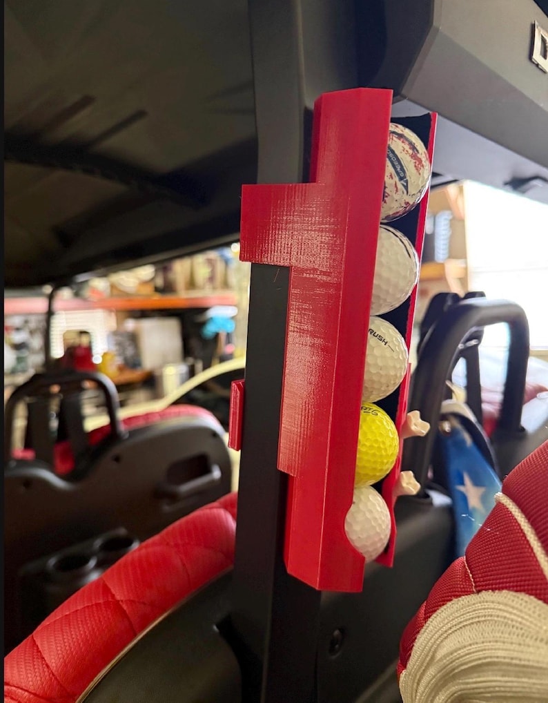 May include: A red golf ball holder attached to a black support post, holding four golf balls. The golf balls are white, yellow, and patterned. The holder is mounted on a golf cart, with a red seat visible in the foreground.