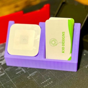 May include: A purple plastic business card holder with two compartments. The left compartment holds a white square object. The right compartment holds a stack of business cards with the text "K3D DESIGNS" printed on them.