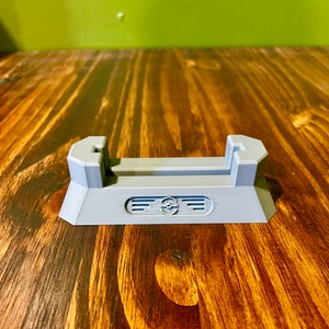 May include: A gray plastic stand with a slot for holding a phone or other device. The stand has a raised base and a decorative design on the front.