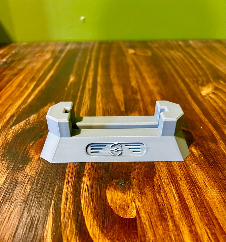 May include: A gray plastic stand with a slot for holding a phone or other device. The stand has a raised base and a decorative design on the front.