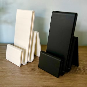 May include: Two phone stands, one white and one black, made of plastic. The stands are designed to hold a phone upright.