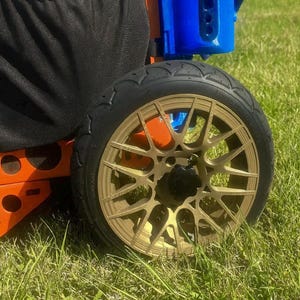May include: Close-up of a black tire with a gold-colored wheel. The wheel has a multi-spoke design and a black center cap. The tire is on a green grassy surface. Orange and blue components are visible.