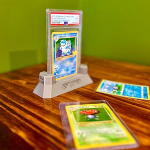 May include: A gray plastic card holder with a clear plastic case holding a Blastoise Pokemon card from the 2021 Pokemon Celebrations Blastoise Holo Classic Collection Base Set.  There are two other Pokemon cards on a wooden surface.