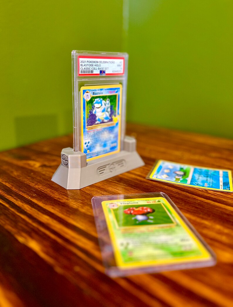 May include: A gray plastic card holder with a clear plastic case holding a Blastoise Pokemon card from the 2021 Pokemon Celebrations Blastoise Holo Classic Collection Base Set.  There are two other Pokemon cards on a wooden surface.
