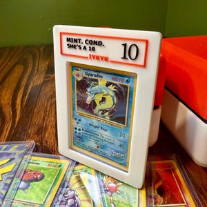 May include: A white 3D-printed display frame holding a Gyarados trading card. The frame features orange text reading MINT. COND. SHE'S A 10 and IYKYK with a large number 10. Additional trading cards in clear protective sleeves are visible below.