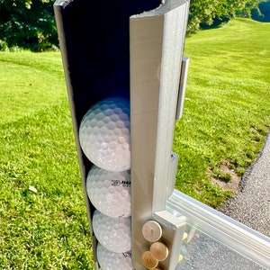 May include: A gray golf ball holder with four white golf balls and several wooden ball markers. The holder is mounted on a golf cart and is set against a backdrop of green grass and trees.