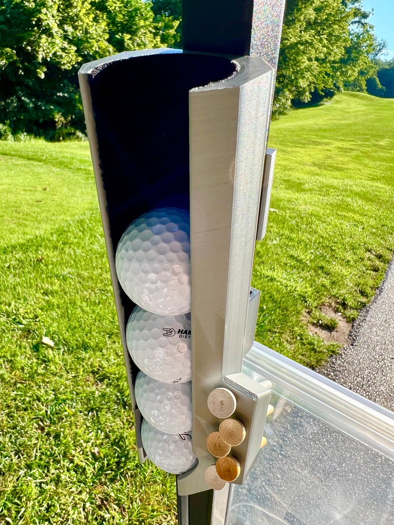May include: A gray golf ball holder with four white golf balls and several wooden ball markers. The holder is mounted on a golf cart and is set against a backdrop of green grass and trees.