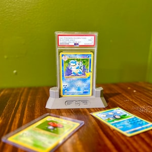 May include: A graded Pokemon Blastoise card from the 2021 Pokemon Celebrations Classic Collection Base Set. The card is in a protective case and is displayed on a stand. The card is graded as a 9, which is considered to be in mint condition.