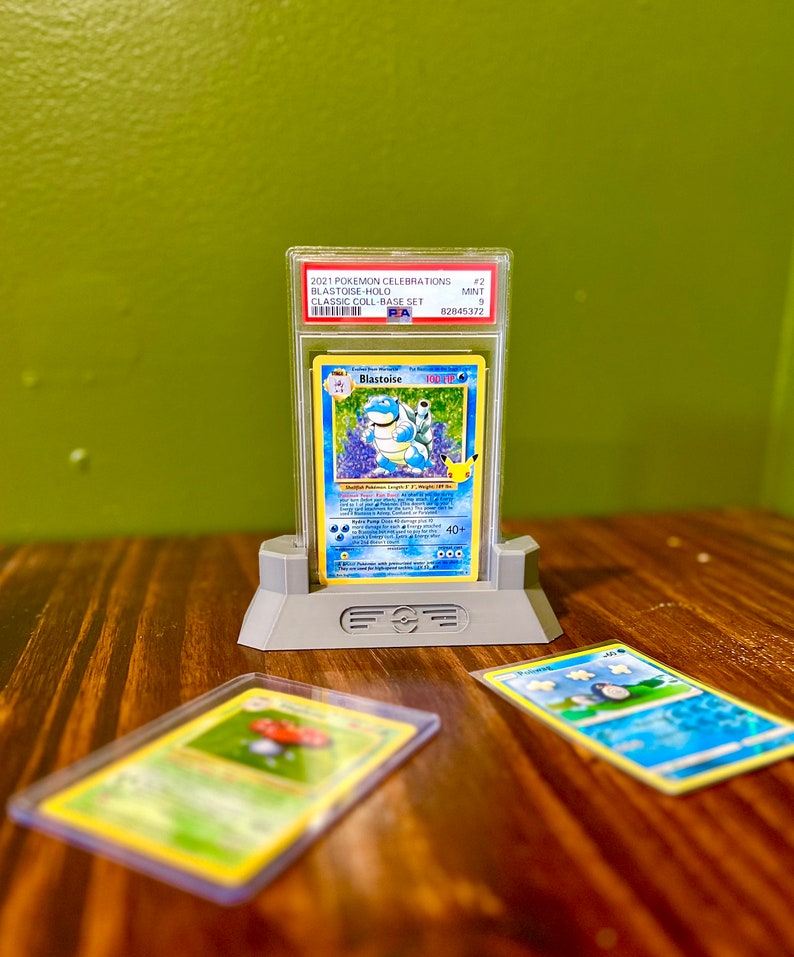 May include: A graded Pokemon Blastoise card from the 2021 Pokemon Celebrations Classic Collection Base Set. The card is in a protective case and is displayed on a stand. The card is graded as a 9, which is considered to be in mint condition.