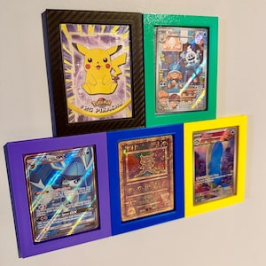 May include: Five framed Pokémon trading cards. The frames are black, green, purple, blue, and yellow. The cards feature various Pokémon characters, including Pikachu, and other characters. The cards are displayed on a white wall.