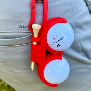 May include: Red golf ball holder with a clip, holding two white golf balls and a wooden tee. The holder has the text "TOP FLITE" and "3" printed on it.