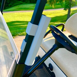 May include: A golf cart interior featuring a black steering wheel and a white and black dashboard. The cart's frame has white and gray accents. The background shows a green golf course.