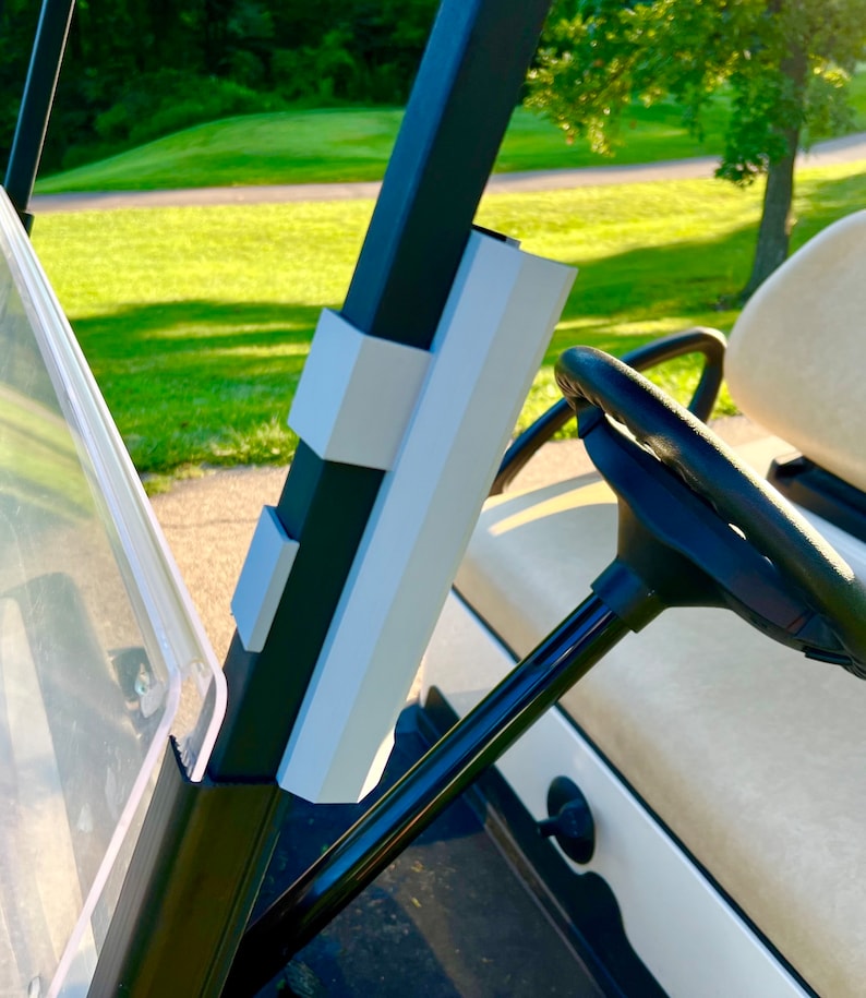 May include: A golf cart interior featuring a black steering wheel and a white and black dashboard. The cart's frame has white and gray accents. The background shows a green golf course.
