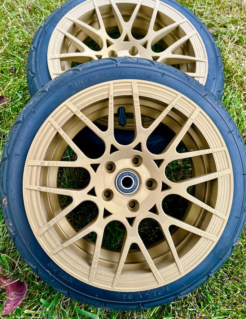 May include: Two gold-colored wheels with black tires. The wheels have a multi-spoke design and are set against a green grassy background. The tires have the text "INFLATE TO 20 P.S.I."