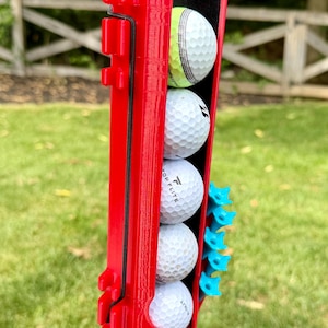May include: A bright red vertical golf ball holder attached to a black post, containing five white golf balls stacked in a column. The holder features a black elastic cord and a side attachment holding five small, light blue plastic golf tees.