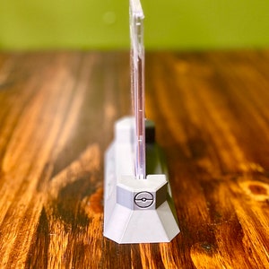 May include: A clear plastic display stand with a gray base featuring a Pokeball logo. The stand is designed to hold a trading card.