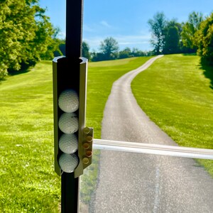 May include: A golf cart accessory holds four white golf balls. The holder is attached to a dark metal frame. The background shows a paved path winding through a green grassy area under a blue sky.