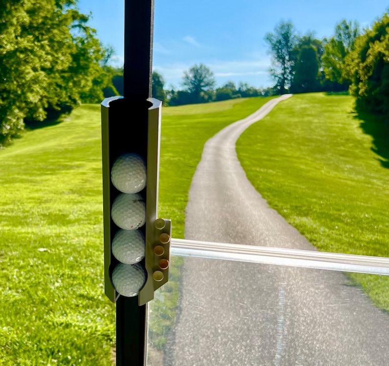 May include: A golf cart accessory holds four white golf balls. The holder is attached to a dark metal frame. The background shows a paved path winding through a green grassy area under a blue sky.