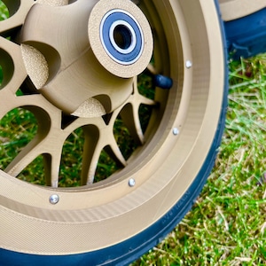 May include: Close-up of a pair of gold-colored motorcycle wheels with black tires. The wheels have a multi-spoke design and visible silver bolts. The tires are mounted on a green grassy surface.
