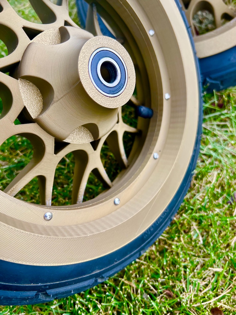 May include: Close-up of a pair of gold-colored motorcycle wheels with black tires. The wheels have a multi-spoke design and visible silver bolts. The tires are mounted on a green grassy surface.