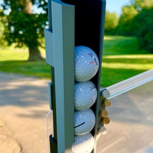 May include: A golf cart accessory featuring a holder for golf balls and tees. The holder is gray and holds three golf balls, each with the number "3" printed on them. Several wooden tees are also visible.