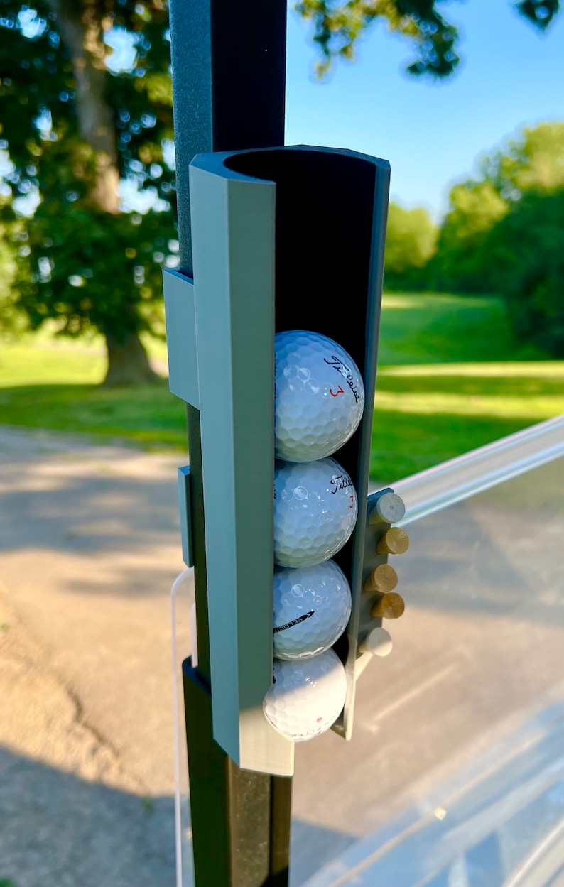 May include: A golf cart accessory featuring a holder for golf balls and tees. The holder is gray and holds three golf balls, each with the number "3" printed on them. Several wooden tees are also visible.
