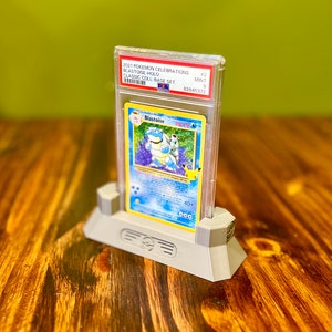 May include: A graded Pokemon trading card of Blastoise from the 2021 Pokemon Celebrations Classic Collection Base Set. The card is in a clear plastic case with a white label that reads "2021 Pokemon Celebrations Blastoise-Holo Classic Coll-Base Set #2 MINT 9 82845372". The card is displayed on a gray plastic stand.