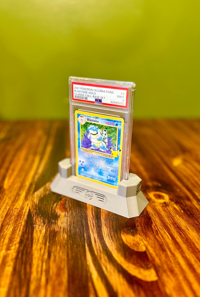 May include: A graded Pokemon trading card of Blastoise from the 2021 Pokemon Celebrations Classic Collection Base Set. The card is in a clear plastic case with a white label that reads "2021 Pokemon Celebrations Blastoise-Holo Classic Coll-Base Set #2 MINT 9 82845372". The card is displayed on a gray plastic stand.