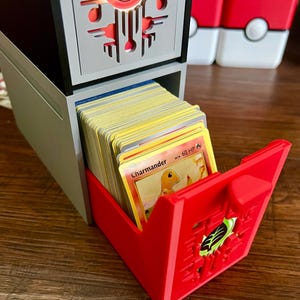 May include: A two-tiered card storage box in gray and black, with a red pull-out drawer. The drawer is filled with trading cards, with a Charmander card visible. The top of the box features a red and black design.