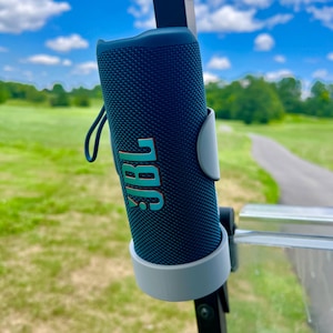 May include: A navy blue cylindrical portable speaker with the word "JBL" in teal and orange. The speaker is mounted on a golf cart, with a green golf course and blue sky in the background.