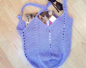 May include: A handmade, lavender crochet tote bag with two handles. The bag is filled with a book and a small card. A pair of sunglasses rests on top. The bag is on a light wood surface.