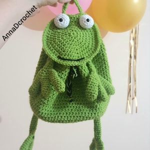以下が含まれることがあります： 大きな白い目と笑顔の口が特徴的な、手作りの緑色のカギ針編みバックパック。カエルを模したデザインで、巾着開閉式とカエルの足が付いています。「AnnaDcrochet」の文字が左側に表示されています。