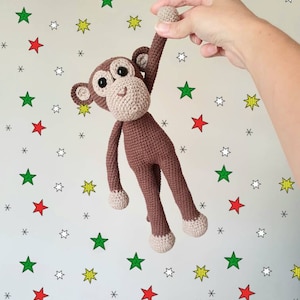 Puede incluir: Un juguete de mono de crochet marrón con una cara sonriente y ojos grandes. El mono está levantando un brazo y está rodeado de un fondo blanco con estrellas rojas, verdes, amarillas y blancas.