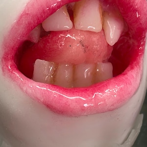 May include: A sculpture of an open mouth with glossy pink lips, white teeth, and a visible tongue. The interior of the mouth is a deep red, and the surrounding skin-like surface is a smooth, matte white material with a realistic texture.