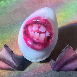 May include: A whimsical, white egg-shaped sculpture featuring a pair of purple duck feet and a detailed, open-mouthed design with pink lips and teeth. The sculpture is set against a backdrop with rainbow hues.