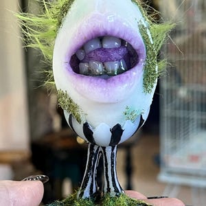 Puede incluir: Una pieza de arte artesanal caprichosa que presenta una cara estilizada con la boca abierta con labios morados y dientes. La cabeza es blanca con detalles verdes y una base a rayas en blanco y negro. La pieza está adornada con material verde similar a la hierba.