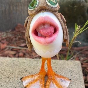 May include: A white egg-shaped figurine with a realistic open mouth showing teeth and a pink tongue. It wears a brown textured aviator-style cap with two round green glass goggles on top. The figure stands on two orange webbed bird feet.