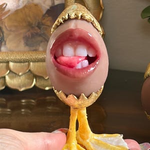 May include: A tan egg-shaped object featuring a realistic human mouth with pink lips, white teeth, and a tongue. The egg is cracked at the top and bottom, with gold-colored edges, and is supported by two yellow, textured webbed bird feet.