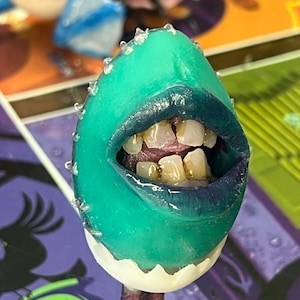 May include: A quirky, artistic egg-shaped sculpture featuring a teal mouth and lips, with visible teeth. The egg has a white base and a purple stem. The design is unusual and striking, with a unique aesthetic.
