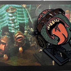 May include: A detailed enamel pin featuring a stylized, open-mouthed creature with a large tongue. The pin has a brown, green, and black color scheme, with a detailed design. The background shows a steampunk-inspired scene with machinery.