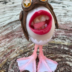 May include: A small figurine featuring a white egg-shaped body with a wide-open mouth showing teeth and a tongue. It wears a brown aviator cap with two amber-colored goggles on top and stands on two pale pink webbed bird-like feet.
