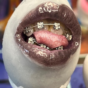 May include: A close-up of a sculpture of a mouth with dark purple lipstick and metal braces. The tongue is pink and extended. The sculpture is white and egg-shaped.