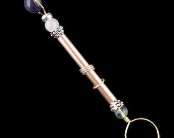 May include: A copper-colored wand with a gold ring at the end. The wand is adorned with silver-colored metal accents and beads in purple, white, and green. The wand is set against a black background.