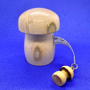 May include: A wooden mushroom-shaped tape measure with a light brown cap and stem. The tape measure extends from the mushroom's side, showing measurements in inches. A smaller mushroom charm hangs from the end of the tape.