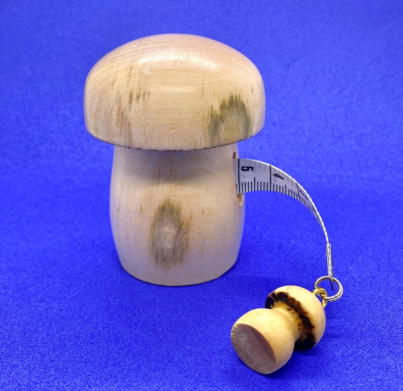 May include: A wooden mushroom-shaped tape measure with a light brown cap and stem. The tape measure extends from the mushroom's side, showing measurements in inches. A smaller mushroom charm hangs from the end of the tape.