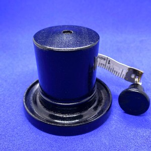 K&ouml;nnte beinhalten: Ein schwarzes, holzfarbenes Objekt in Zylinderhutform mit einem kleinen Loch in der Mitte, das auf einer kreisf&ouml;rmigen Basis sitzt. Ein kleiner schwarzer Knopf und ein wei&szlig;es Ma&szlig;band befinden sich neben dem Hut. Das Band zeigt "109 L".