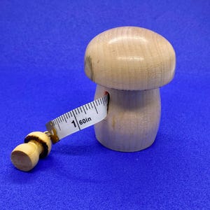 May include: A wooden mushroom-shaped measuring tape holder with a white measuring tape extending from its side. The tape reads "1 60in". The mushroom is light brown and sits on a blue background. The tape is 60 inches long.