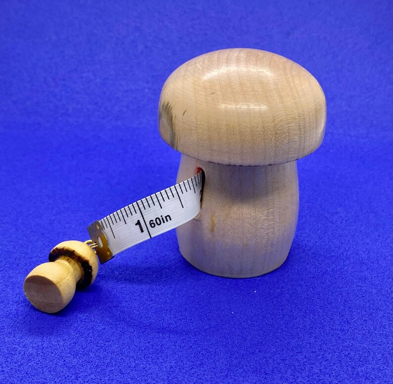 May include: A wooden mushroom-shaped measuring tape holder with a white measuring tape extending from its side. The tape reads "1 60in". The mushroom is light brown and sits on a blue background. The tape is 60 inches long.